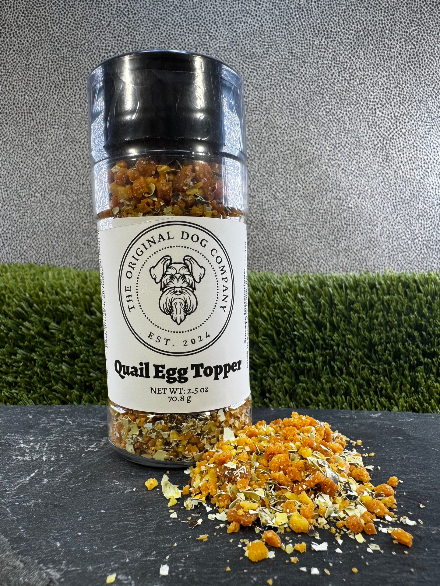 Bottle of 'Quail Egg Topper' from The Original Dog Company on a dark surface with grass in the background.