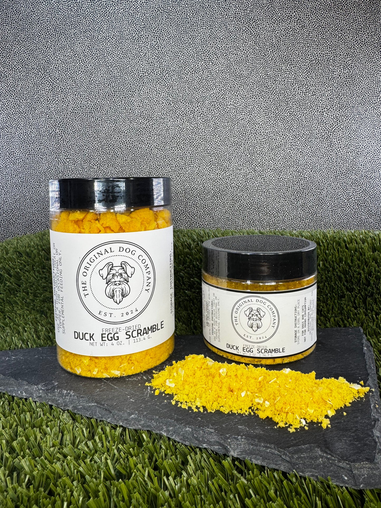 Two jars of 'Duck Egg Scramble' from The Original Dog Company on a grassy surface with a gray background.