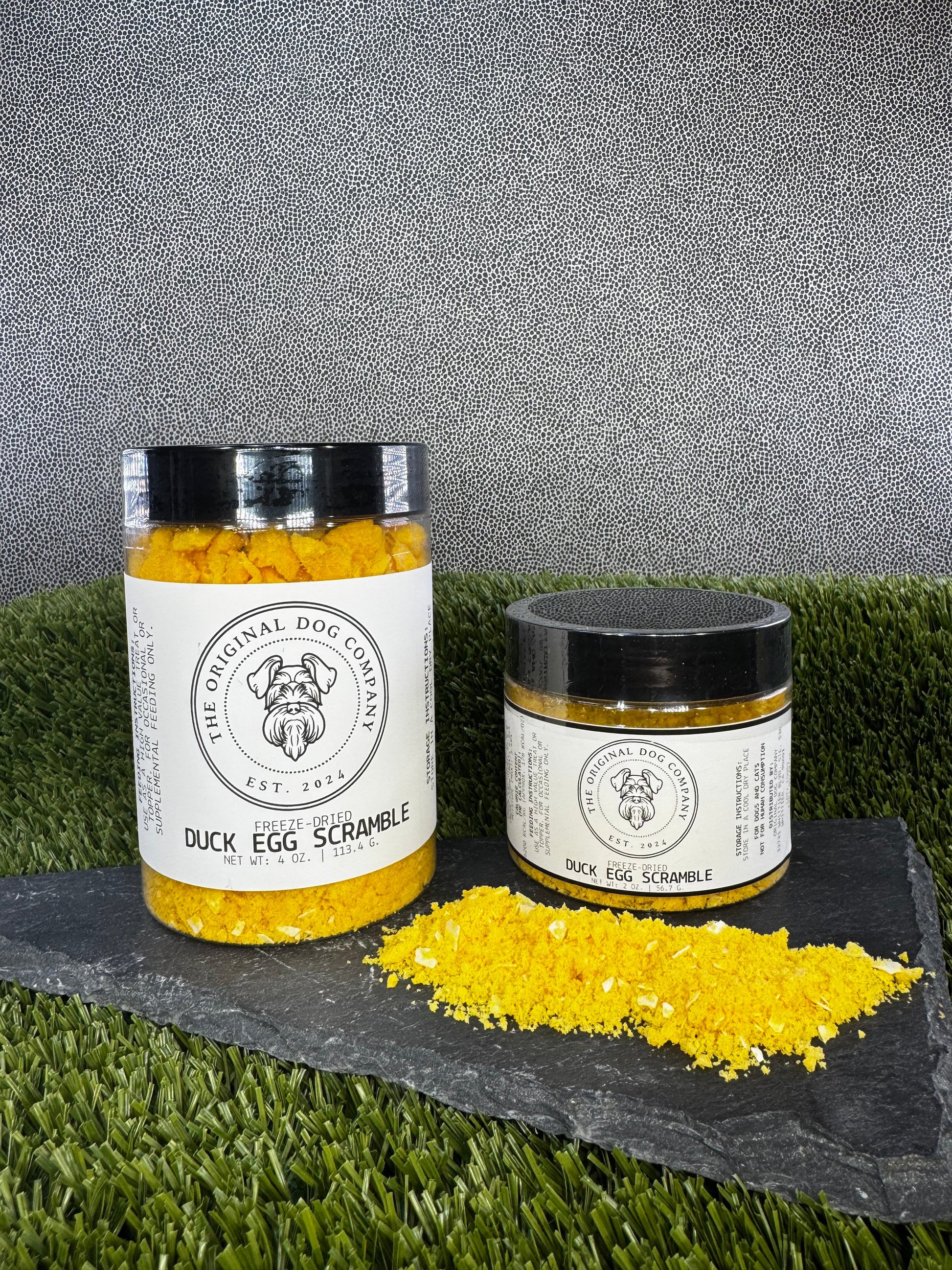 Two jars of 'Duck Egg Scramble' from The Original Dog Company on a grassy surface with a gray background.